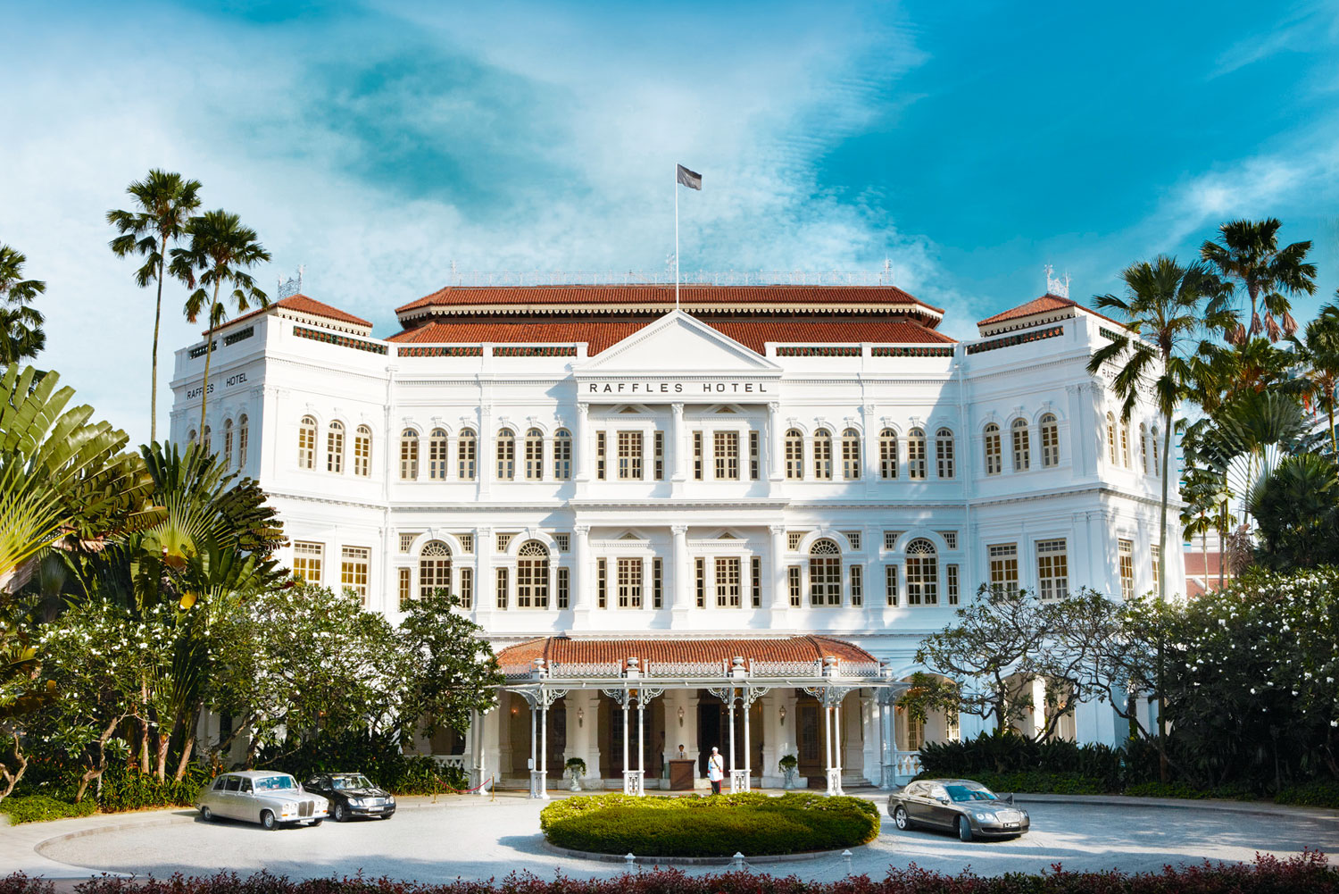 Front view of historic Raffles Hotel with vintage cars parked outside and tropical landscaping