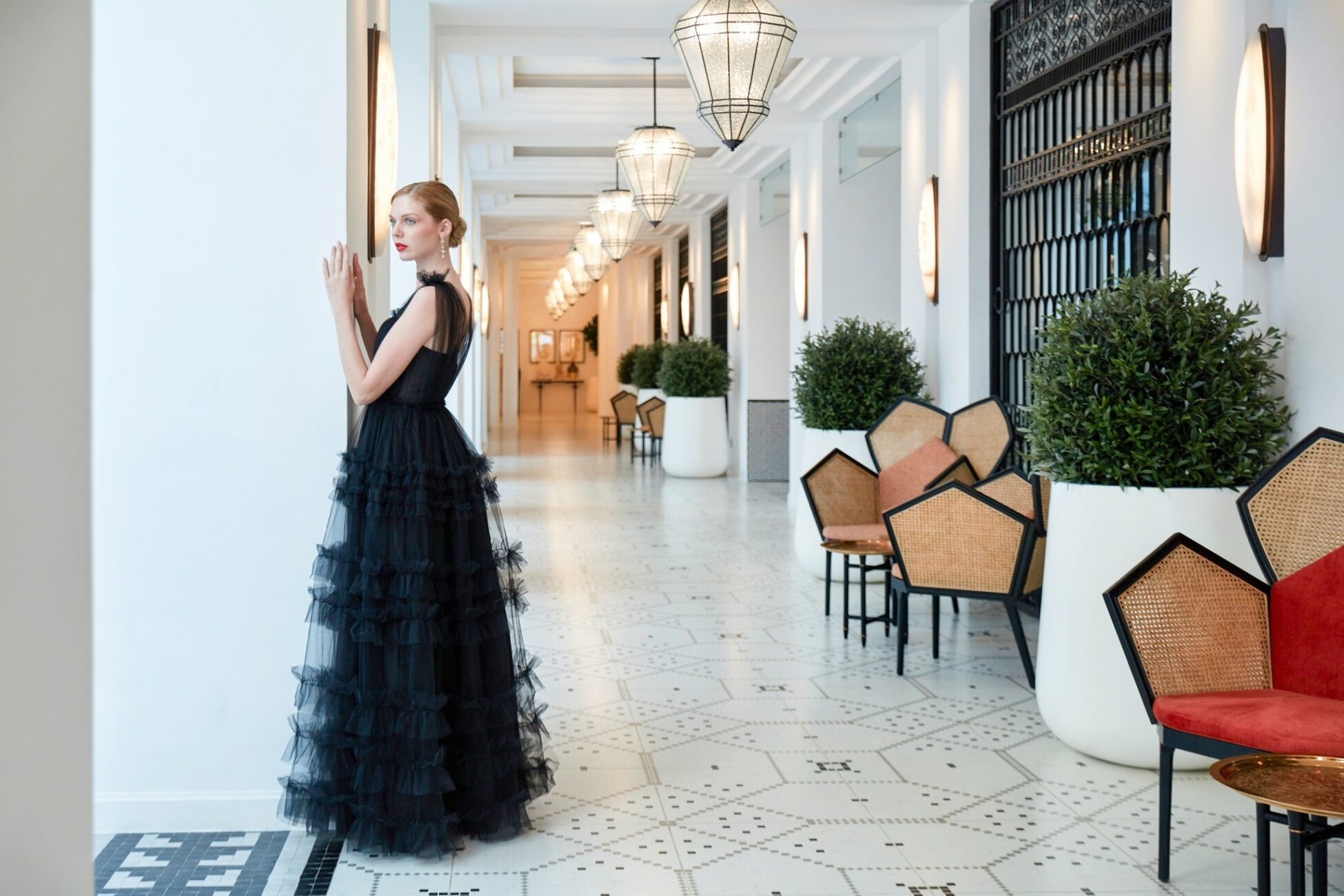 Elegant woman in long black ruffled gown standing in bright hallway with patterned tile floor and modern decor