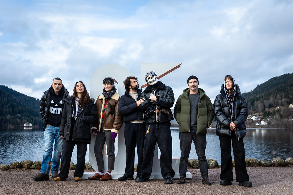 Group of seven people dressed in winter clothes standing outdoors by a lake with mountains in background