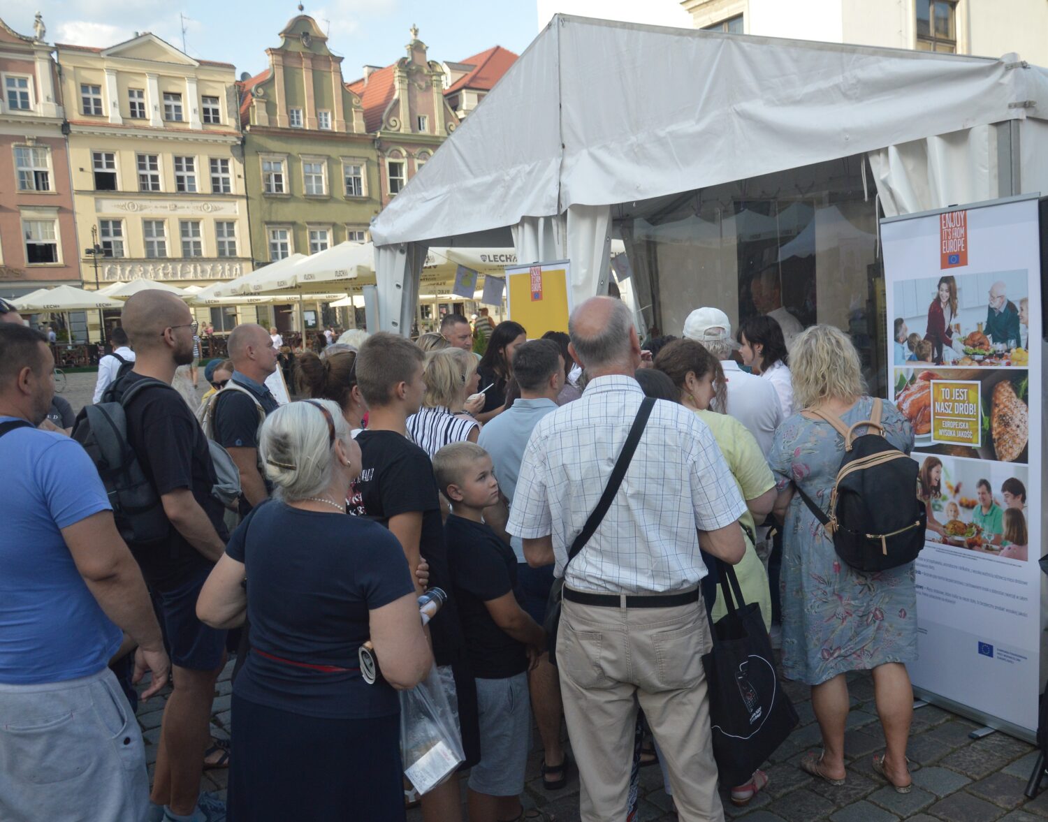 Crowd gathered outside a white tent in a historic city square watching a cooking demo with “Enjoy It’s From Europe – To jest nasz drób” banner
