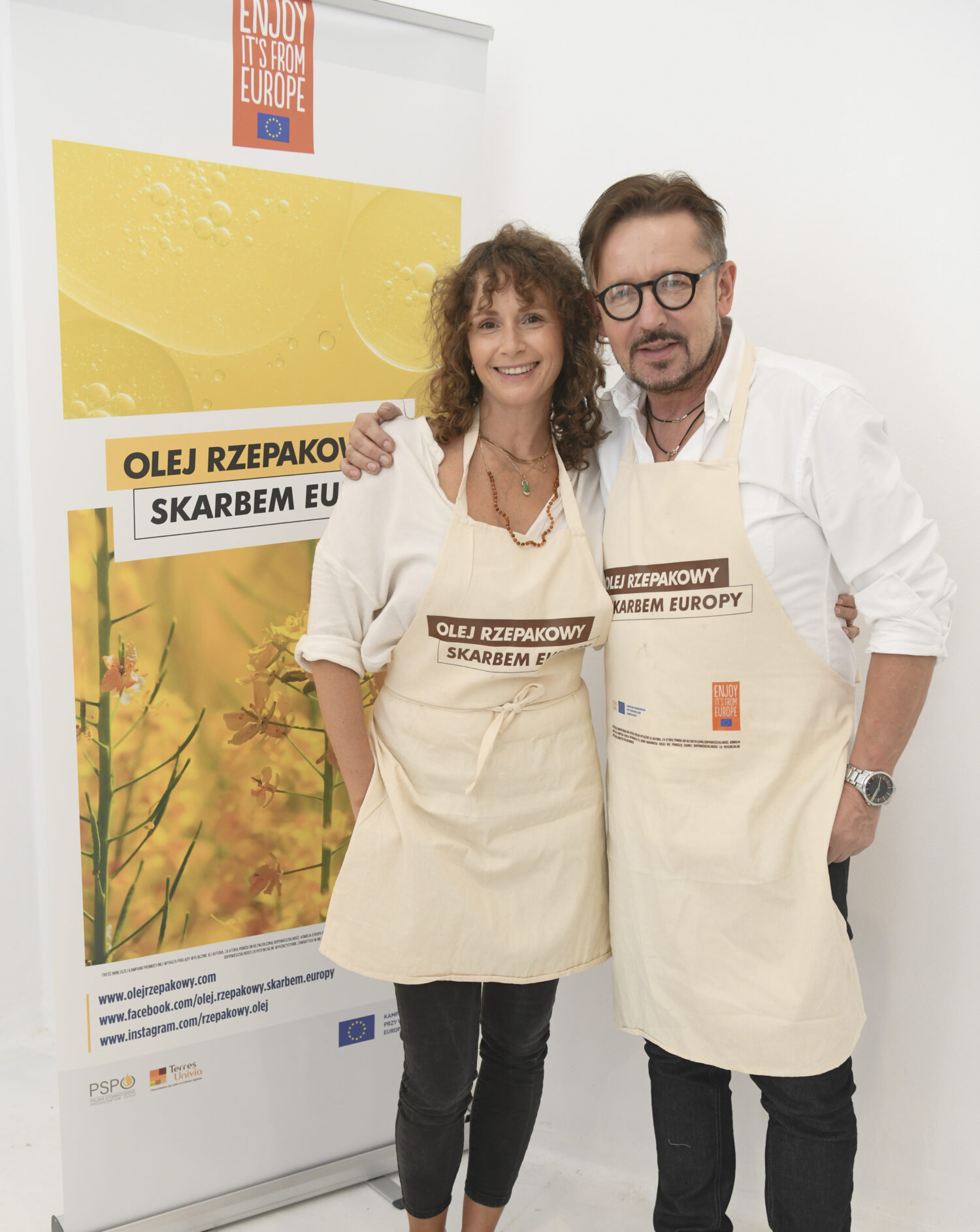 Two people in cream aprons posing in front of “Enjoy It’s From Europe – Olej rzepakowy skarbem Europy” rapeseed oil campaign banner